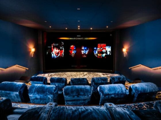 Zdjęcie przedstawiające 10 niebieskich foteli w dwóch rzędach przed ekranem, na którym widnieją cztery twarze. Przedstawiona instalacja to pierwsze europejskie domowe kino typu IMAX. Zdjęcie pochodzi ze strony https://www.cepro.com/