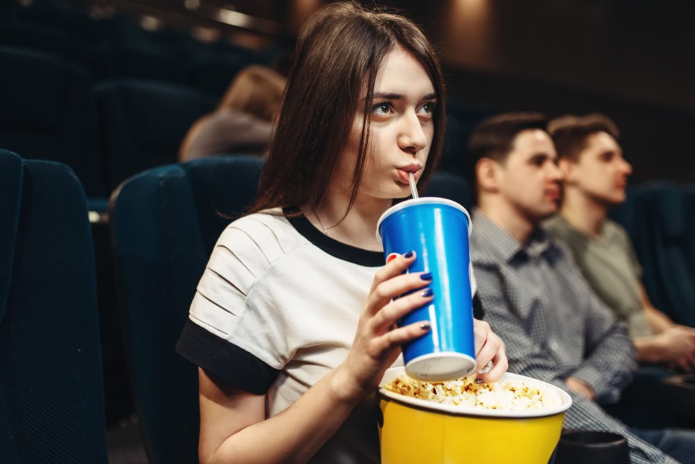 Zdjęcie przedstawiające dziewczynę pijącą napój i jedzącą popcorn na sali kinowej